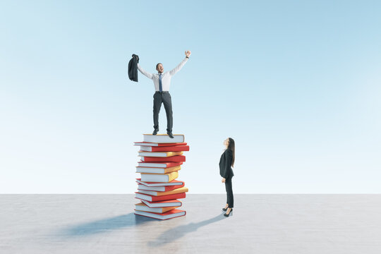 Young european businesswoman looking up at happy colleague standing on book pile. Blue sky background. Education, knowledge and success concept.