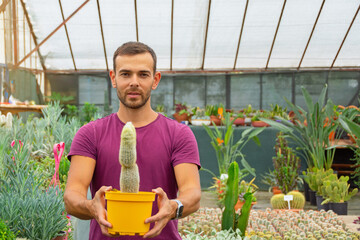 The guy swarthy gardener holds a pot with a fluffy hairy cactus grown in a greenhouse.