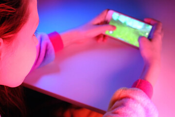 Closeup portrait of pretty young girl playing online mobile game on a smart phone. Teen girl play the game on cellphone. Gamer playing online game on smartphone in dark room. Neon blue and pink light.