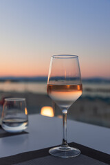 The pink glow of a sunset along with the sea and sandy beach is reflected in a focused glass of white wine on a table in a beach cafe