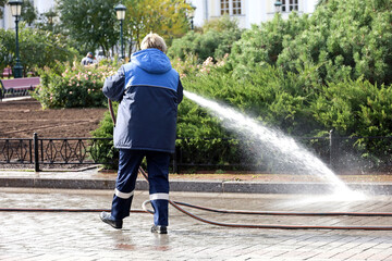 Woman worker in blue uniform watering the sidewalk with a hose. Street cleaning and disinfection in...