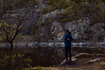 A man with a backpack standing in a forest on a rock next to a lake at sunrise.