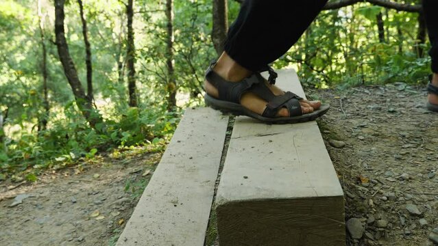 Men's feet in black pants and black trekking sandals climbing a wooden ladder in the woods. Side view. Close-up. Slow motion. Active recreation in nature
