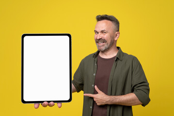 Cheerful bristle and mustached senior man dressed in green shirt and brown t-shirt pointing with thumb up and finger to his big tablet pc with white mock-up screen holding in hand on yellow background
