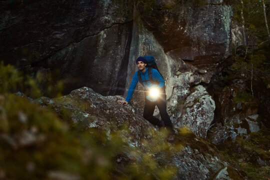 Adventurous male hiker exploring a dark rocky cave at night, holding a bright flashlight and wearing a blue jacket and backpack. High-quality outdoor exploration and wilderness survival concept.