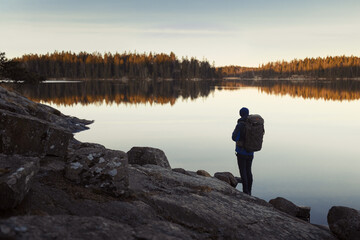 Fototapeta na wymiar A caucasian man with a backpack standing on a rock in a forest next to a lake at sunrise.