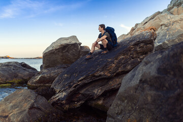 A caucasian man sitting on a rock by the sea with backpack.