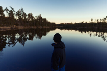 A man standing by a lake in the forest at sunset watching a blue sky reflecting in the water.