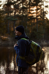 A caucasian man standing by a lake in a forest with a backpack at sunset.