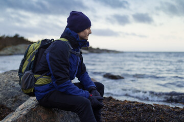 A caucasian man sitting on a rock by the coast with a backpack looking out over the sea.