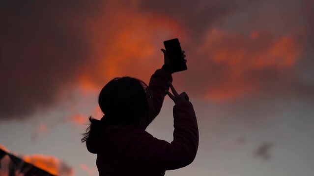 Silhouette of Indian female holding mobile phone and posing for selfie in background of sunset sky. Young Indian Travel Woman Vlogger vlogging during sunset in mountains.  