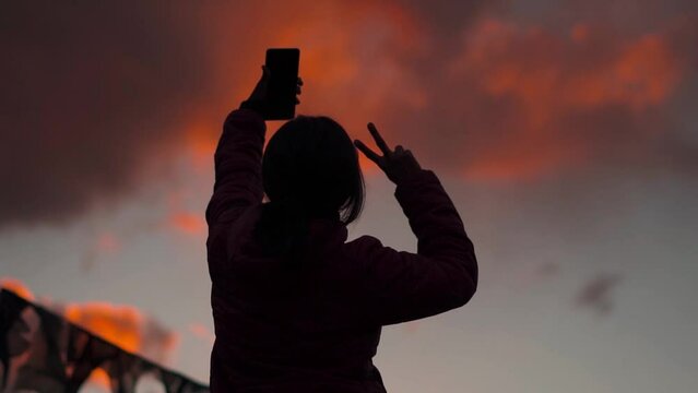 Silhouette of Indian female holding mobile phone and posing for selfie in background of sunset sky. Young Indian Travel Woman Vlogger vlogging during sunset in mountains. Holidays vacation concept