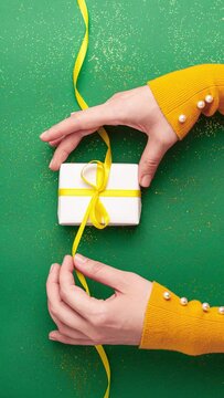 Human hands tied a yellow satin ribbon bow on white gift box on jade green background with gold colored glitter. Stop motion vertical animation Christmas Holidays and present concept flat lay