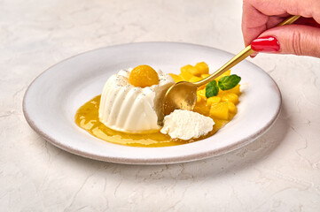 Woman's hand with spoon breaks off piece of homemade panna cotta with natural vanilla pod, mango syrup and mint, selective focus
