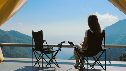 Back view of woman sitting in folding camp chair, chill and enjoy mountain view near camping tent...
