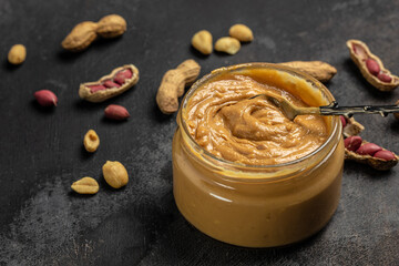 Peanut butter or paste in an open jar and peanuts in the peel scattered on a dark background, banner, menu, recipe place for text, top view