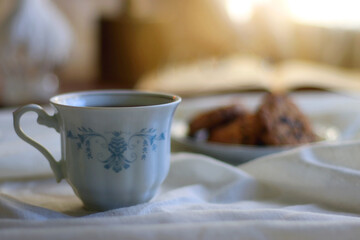 Cup of tea or coffee, plate of chocolate chip cookies, open book, reading glasses, candle and vase with flowers. Selective focus.