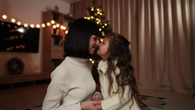 Happy family celebrating Christmas. Mom and cute daughter in warm knitted white sweaters sitting on the background of decorated tree and touching their noses.