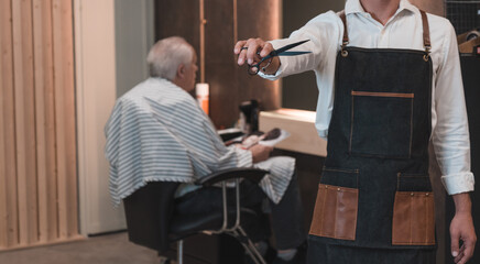 Peluquero irreconocible, mostrando las tijeras, y de fondo cliente senior sentado con la capa, listo para cortar el pelo. - obrazy, fototapety, plakaty