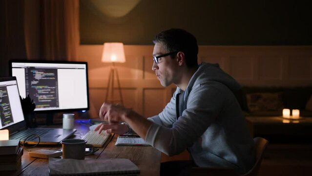 young man in glasses remote works on computers, sits at table at home in darkness. bites apple, types on keyboard looking on monitor intently. hardworking and concentrated on programming due deadline