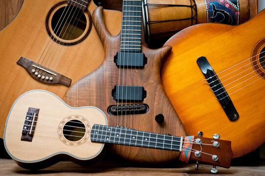 Acoustic and electric guitars, ukulele and djembe standing side by side on a wooden background.