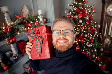 Obraz na płótnie Canvas Happy young man make selfie photo with red gift box on background Christmas interior house