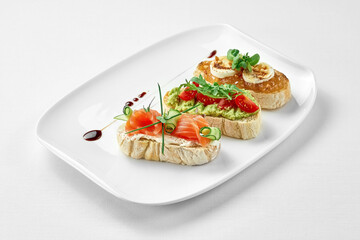 Set of bruschetta with salmon, avocado and goat cheese in a white plate on a white background. Close-up, selective focus.