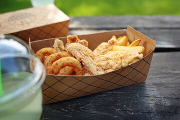 Deep fried onion rings and breaded chicken strips, served with fries, in a takeaway box. Fast food, food truck meal.
