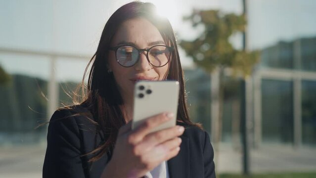 At sunlight smiling beautiful young businesswoman with glasses using a modern smartphone stand in the city smiling. Slow motion