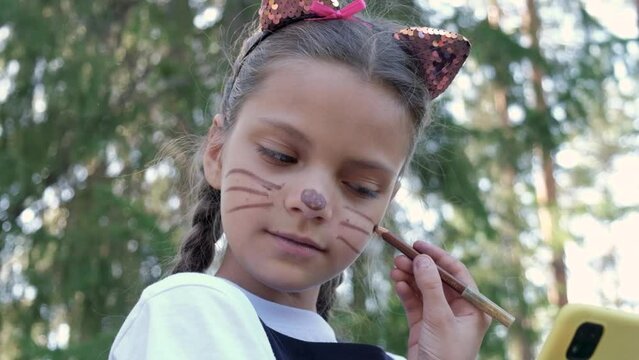 a teenage girl draws a make-up on her face, the image of a cat for halloween, preparation for the holiday of halloween, cat ears on a headband and a painted mustache and nose of a cat on a face