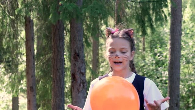 a cheerful girl with a make-up on her face plays with a balloon, the image of a cat for halloween, preparation for the halloween holiday, cat ears on a headband and a painted mustache and nose of a ca
