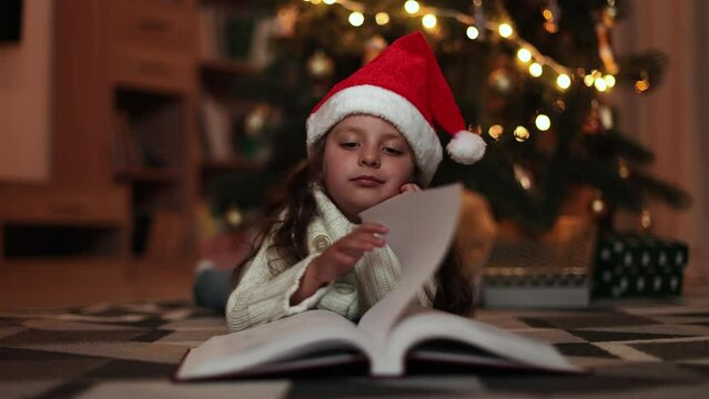Cheerful little preschool girl in knitted warm sweater reading magical fairy tale book lying on floor near Christmas tree at decorated room, merry Christmas.