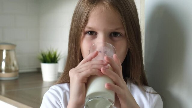 Cute kid girl drinking milk from glass in kitchen at home. Closeup child drinks cup of yogurt or kefir, smiling and looking at camera indoors. Healthy eating for children