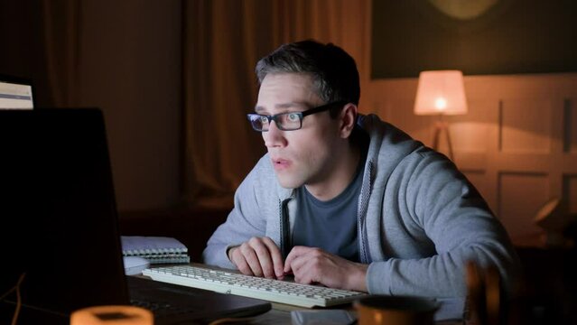male freelancer in eyeglasses remote works at home on laptop at night darkness. programmer gawp intently on monitor with whole wide eyes. he looks worried and nervous, dissatisfied