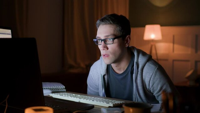 male freelancer in eyeglasses remote works at home on laptop at night darkness. programmer gawp intently on monitor with whole wide eyes. he looks worried and nervous, dissatisfied