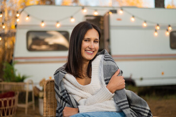 Caucasian woman sits in a wicker chair wrapped in a blanket in the yard near the trailer in autumn. 
