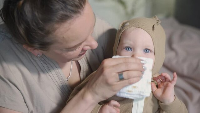 Baby with running nose getting wiped by mother's hand before sleep. Cleaning nose rhinitis. Child is in pajamas. Daily hygiene, parenthood and mother's routine, seasonal cold, flu. Close-up