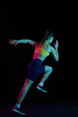 Fototapeta na wymiar Professional female athlete running isolated on dark background in neon light. Healthy lifestyle, skills, sport, fitness, speed and energy concept.