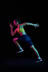 Fototapeta premium Dynamic portrait of young sportive woman training, running isolated on dark background in neon light. Competition, sport, fitness, speed and energy concept.