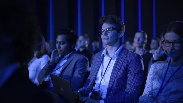 Young Man Sitting in a Crowded Audience at an IT Conference. Delegate Using Laptop Computer to Take Notes. Software Developer Watching Presentation About New High Tech Products and Cloud Solutions.