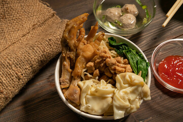 Chicken noodles in white bowl and wooden chopsticks on natural background with chicken pieces, chicken feet, green mustard, meatballs, boiled dumplings, sauce. Indonesian Chicken Noodles