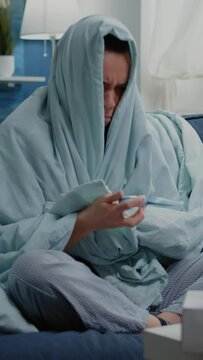 Vertical video: Person wrapped in blanket using thermometer for temperature measurement at home. Ill woman checking fever while feeling cold and shivering. Adult having chills and virus symptoms.