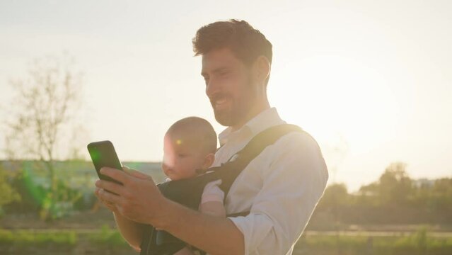 Portrait of happy father with a little newborn baby in a slings carrier. Use phone smiling on street. Ergo backpack. Parents. Gadget. Smartphone. Cell. Slow motion