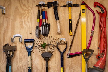 Assortment of DIY gardening tools and equipment hanging organised on wooden wall inside garden shed. 