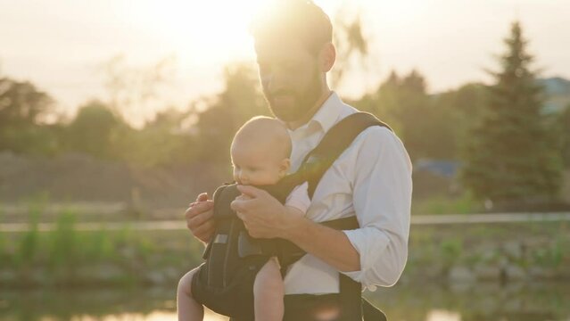 Near lake portrait of funny newborn baby in a slings carrier. Young father holding a child kiss and hug on street at sunlight. Ergo backpack. Parents. Slow motion