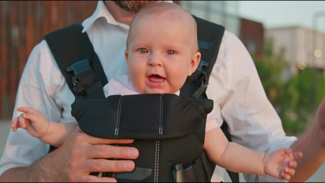 Charming funny newborn baby in a slings carrier wants to sleep and yawns. Young father holding a child on street. Ergo backpack. Parents. Slow motion