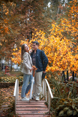 Fototapeta premium A young beautiful couple in love dressed in casual style in an autumn park stands on a bridge, golden autumn