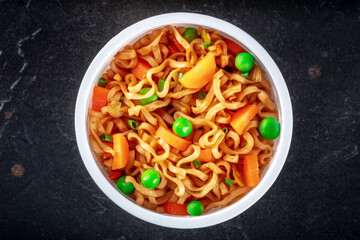 Ramen cup, instant soba noodles in a plastic cup with vegetables, green peas and carrots, top close-up shot on a black slate background