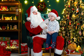 Santa Clause playing with little cute boy catching his nose near Christmas tree. New Year concept 