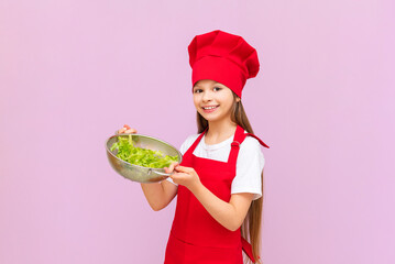 a smiling girl in a red chef's suit holds a sieve with a fresh green salad in her hands.